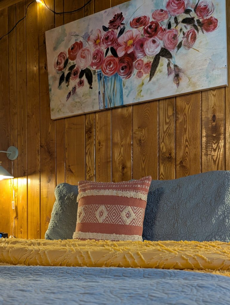 Head board wall of bed that is made with bright color bedding and flower painting hanging on the wall